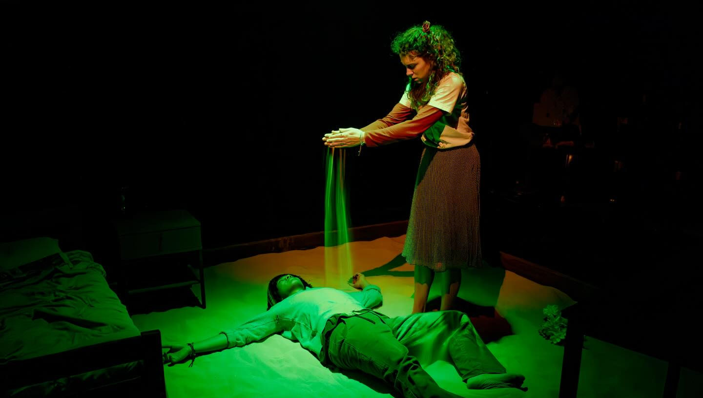 A woman in a white shirt and grey skirt stands, pouring sand over a man in a grey shirt laying in the sand. The man is lit in green, the woman in warm white.