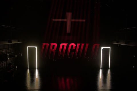 A large projected image of the text “DRACULA” in red on a black wall. Two entrances lined with white light are on either side. Hung above is a crucifix light box that is off.