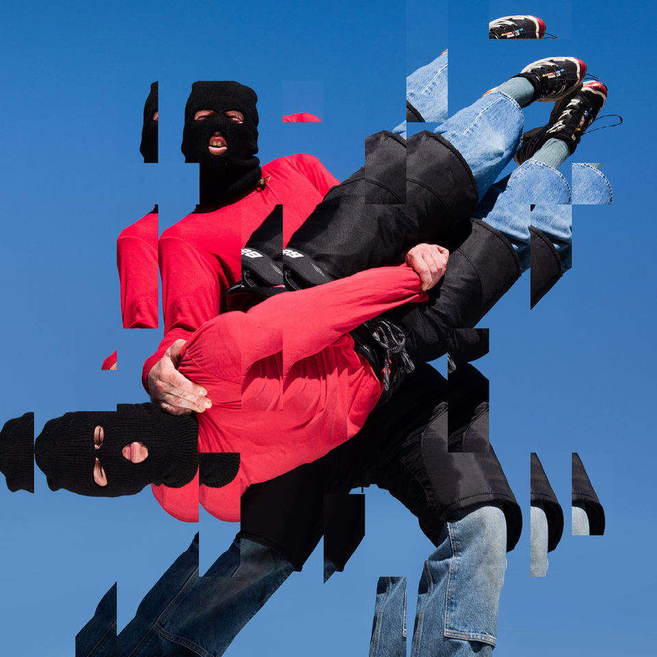 A glitchy photo of a performer in a balaclava, red jumper and jeans, holding another performer, dressed identically, upside down on a blue background.