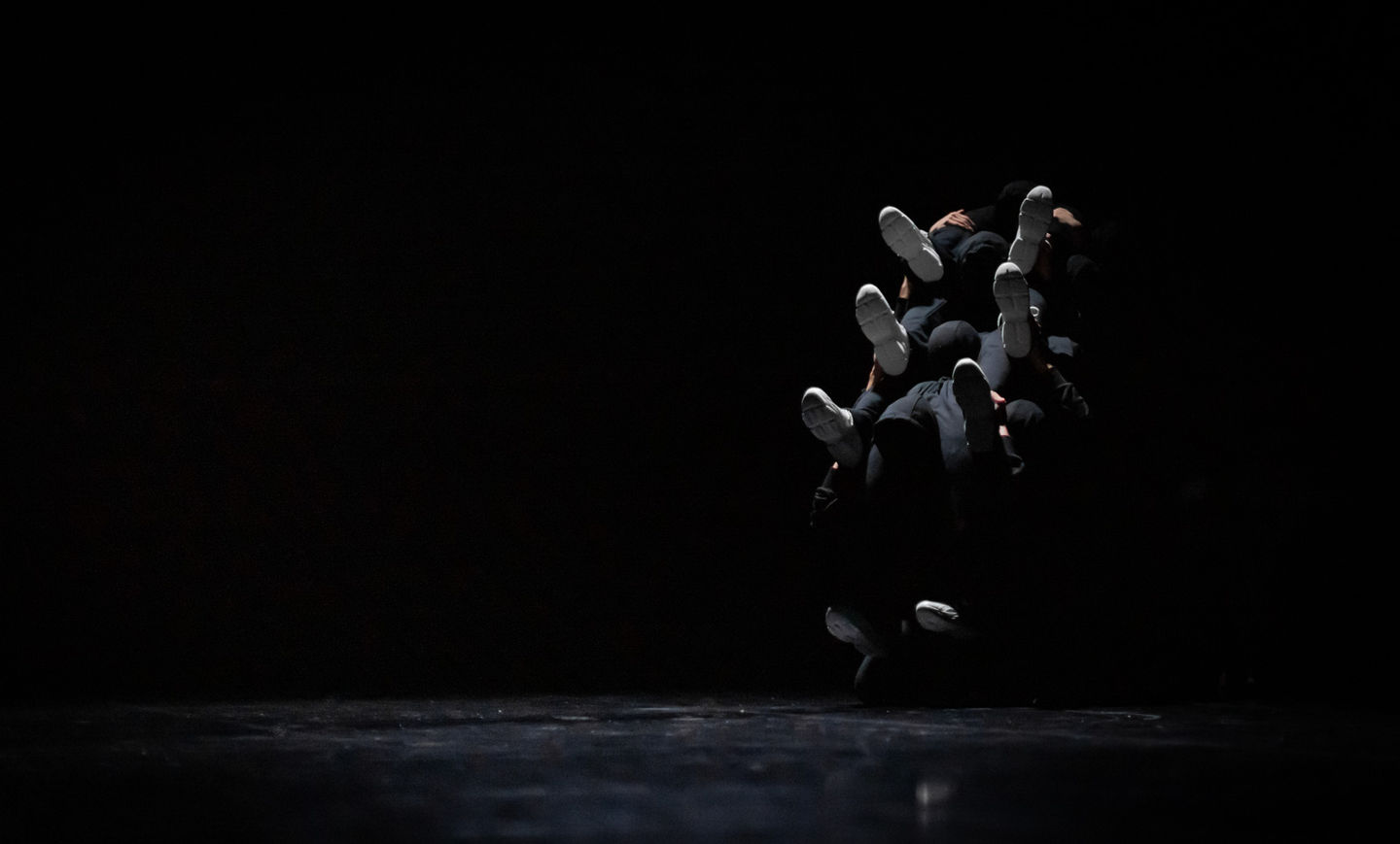 Several dances dressed in black forming a centipede-like mass. They are lit in cold white.