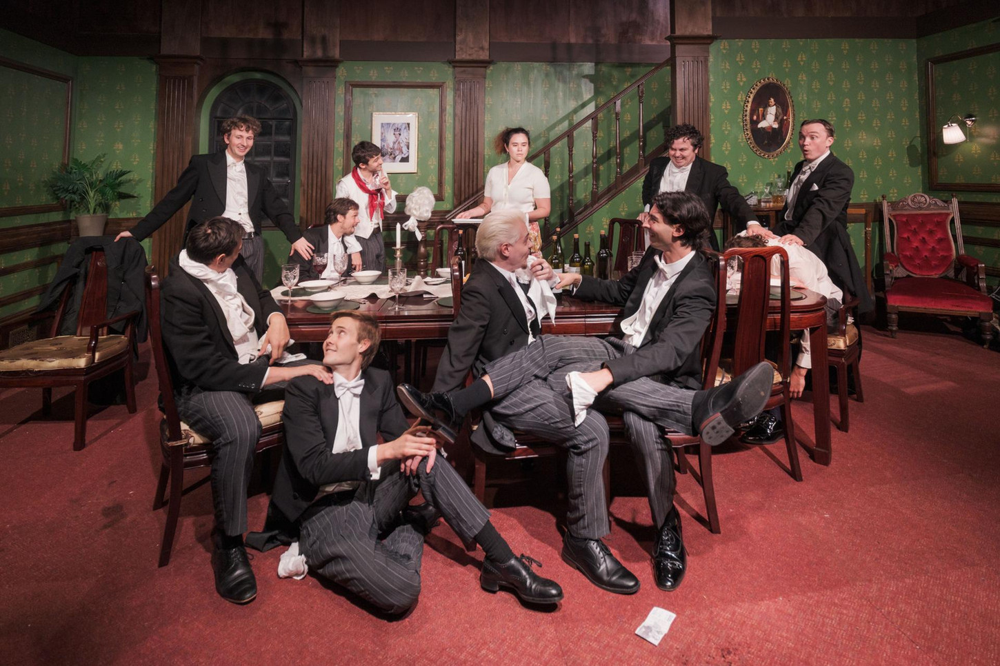 Ten men dressed in full tails are seated around an elegant dining table. In the centre, a woman dressed in a white top and floral skirt, holding a stack of plates. They are lit in warm white.
