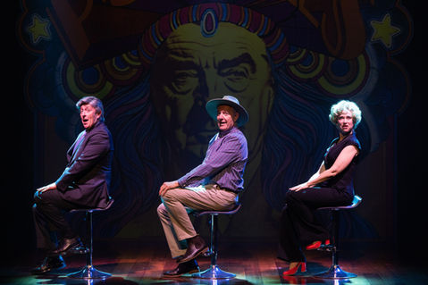 Three performers dressed as politicians seated on stools. On the left, a man in a dark blue suit and grey wig, the centre, a man in brown pants, purple button up and a wide brim hat, the right, a woman in a black one-piece and a blonde curly wig. A large projected image of the Follies poster with Robert Menzies’s face swapped in is visible in the background. They are lit in cool white.