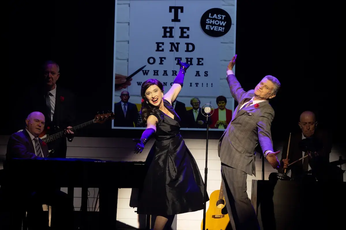 A woman in a black dress and a man in a grey suit stand at a 1950s microphone. Behind them are a man seated at a piano, a man standing, playing the bass and a man sitting at a drum kit, all in suits. A large projected image of the poster for this show is visible in the background. The two in the foreground are lit in cold white, the others are in shadow.