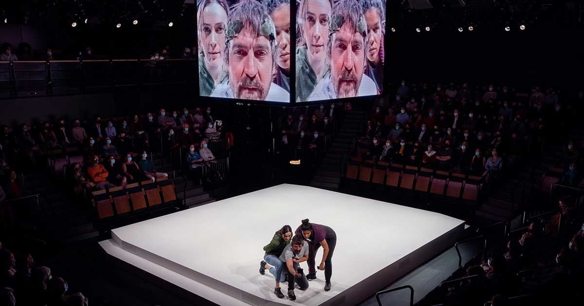 A man and two women stand in the corner of a white square, huddle around a phone. Hung above, a digital screen cube displaying the live feed from that phone's camera. They are all lit in cold white.