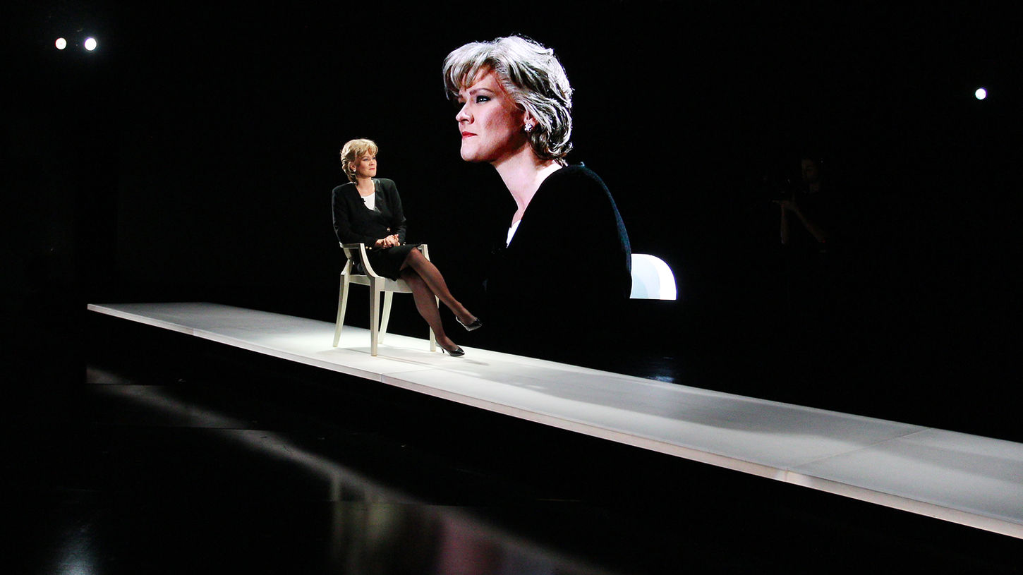 A woman in a black suit sitting in a white chair on a raised white platform, looking out. A large digital screen displaying the live feed of the woman from the opposite angle is visible in the background. The woman is lit in cold white.