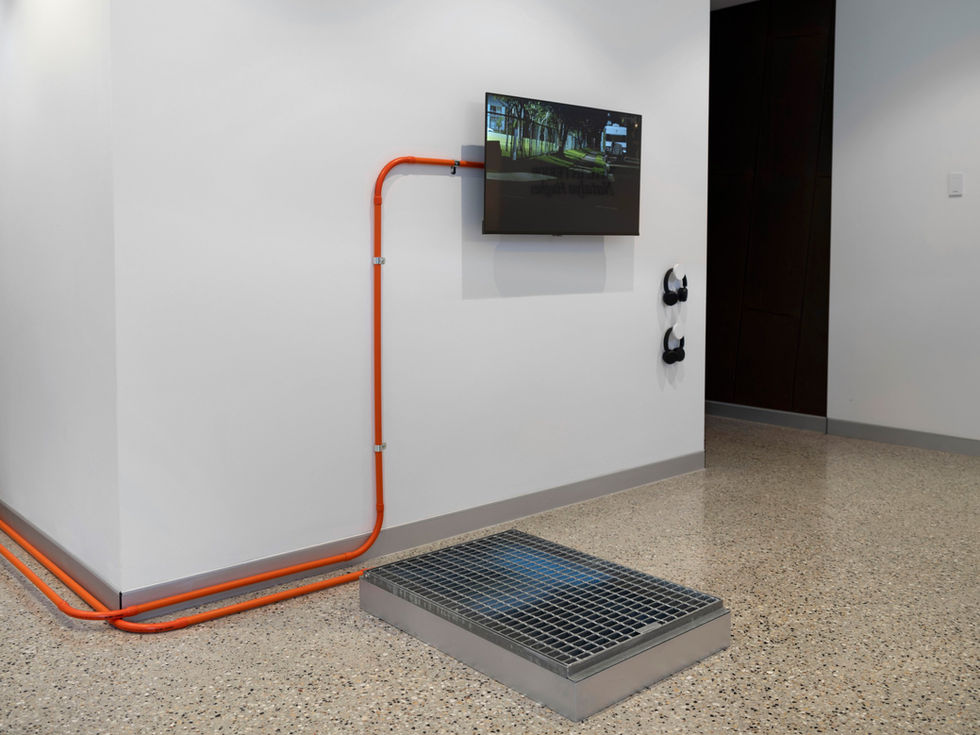 Amanda Bennetts, Latency + Lacerations, 2025. A wall-mounted screen fed by bright orange conduit running along the floor, with a steel floor grate below and headphones on the wall. UniSC Art Gallery, 2026.