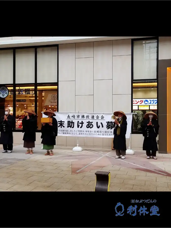 長崎市新大工商店街にて、長崎市仏教連合会の旗を掲げて歳末助け合い募金を呼びかける僧侶。旗には「長崎市仏教連合会歳末助け合い募金」の文字が記されている。