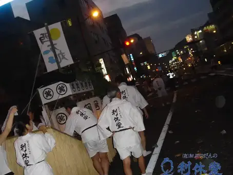 長崎の精霊流しの様子。夜の街路で、お揃いの白い法被の参加者がが昔ながらの藁を利用したちょ木製の精霊船を曳いている。家紋入り提灯や西方丸の旗がある。