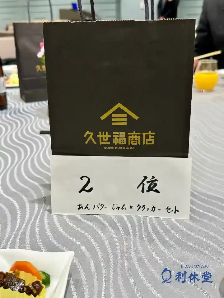 懇親会のテーブル席に置かれた、格付けチェックゲーム2位の景品袋。中には久世福商店の「あんバタージャム」と「クラッカー」のセットが見える。