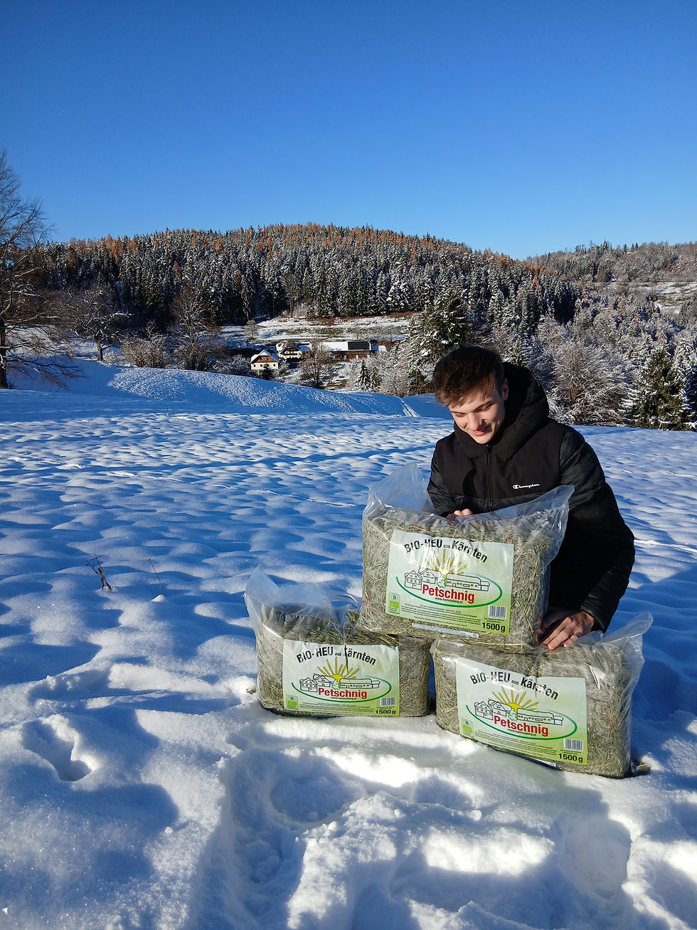 Simon Petschnig im Schnee mit Heu-Paketen, Bio-Heu 1500g, verschneite Landschaft, blauer Himmel.