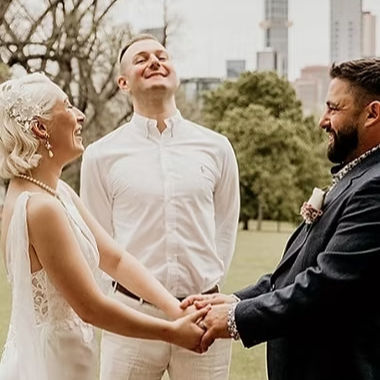 A happy marriage celebrant in Melbourne stands between a laughing bride and groom during an outdoor wedding ceremony.