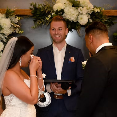 A smiling marriage celebrant in Melbourne stands between a bride, who is wiping away tears, and a groom during a wedding ceremony.