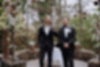 A Melbourne marriage celebrant addresses the crowd at a wedding ceremony, standing beside the groom in a decorative outdoor setting.