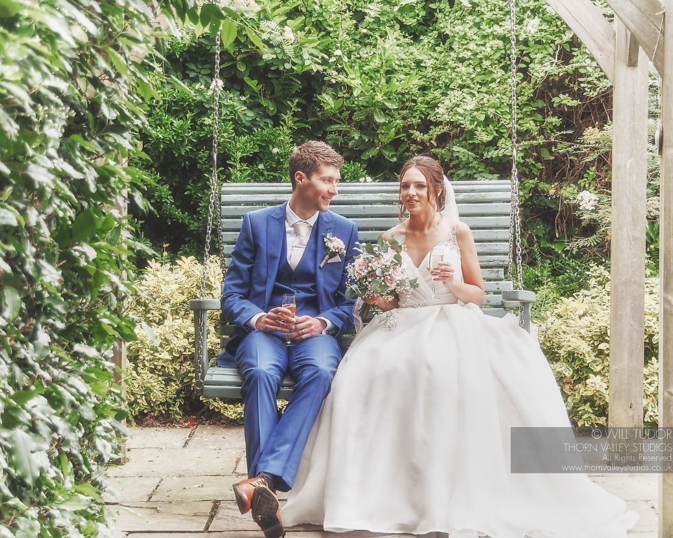 Bride in white gown and groom in blue suit sit on a garden swing, smiling with drinks and bouquet; wedding watermark in corner.