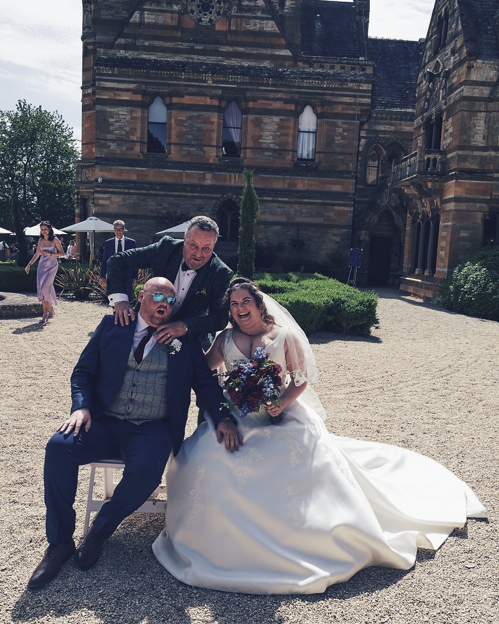 Bride and groom laugh while a man poses behind them outside a historic stone building at a wedding.