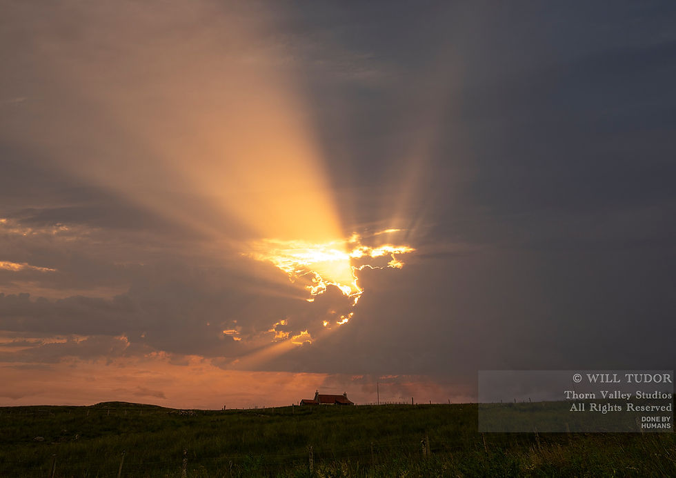 Dramatic sunset rays break through dark clouds over a rural field and small house, with © Will Tudor watermark.