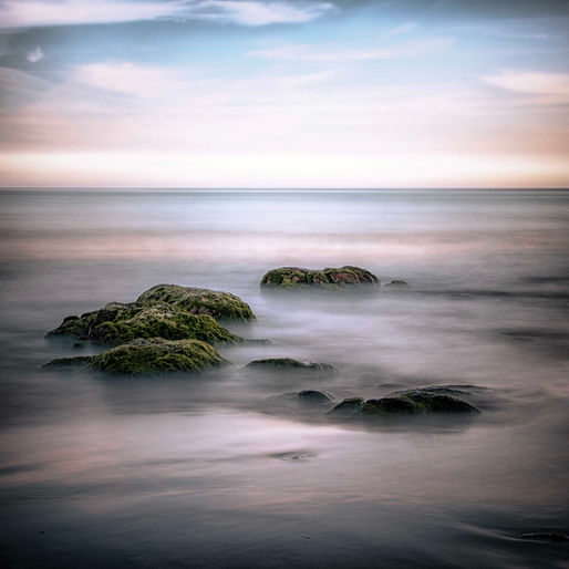 Moss-covered rocks in a calm, long-exposure sea under a pastel sunset sky, creating a serene seascape