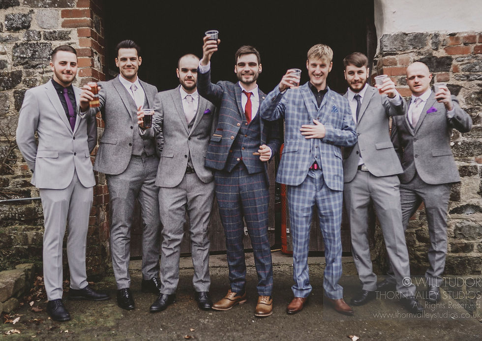 Seven men in suits toast with drinks outside a stone doorway, smiling; Thorn Valley Studios watermark visible.