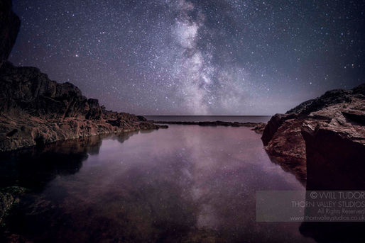 Milky Way glows over rocky coast and still water at night, with dark cliffs framing a purple star-filled sky and watermark text.