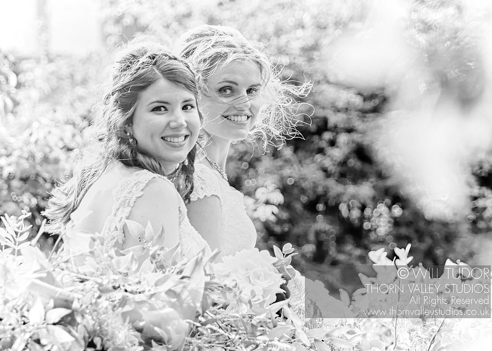 black and white image of two brides facing the camera, smiling through trees on a sunny day