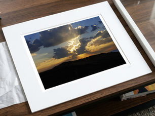 Framed sunset landscape photo on a wooden desk, with dramatic clouds and golden light beside another white frame.