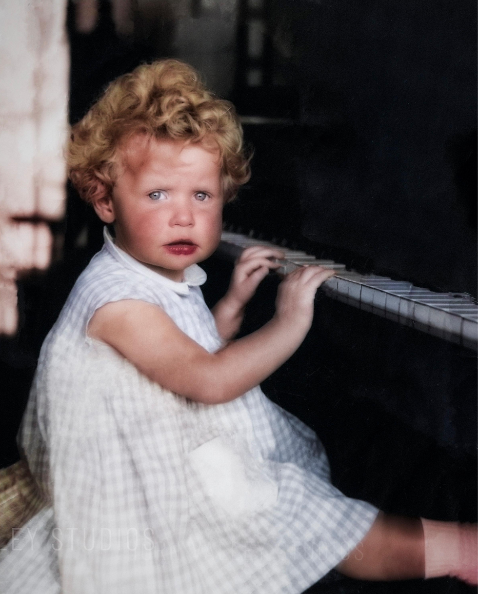 Curly blond toddler in a white checkered dress touches piano keys in a dim room, looking solemn and curious.