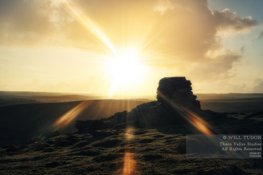 Golden sunrise over rocky moorland, bright sunburst behind a silhouetted rock; watermark text in lower right.