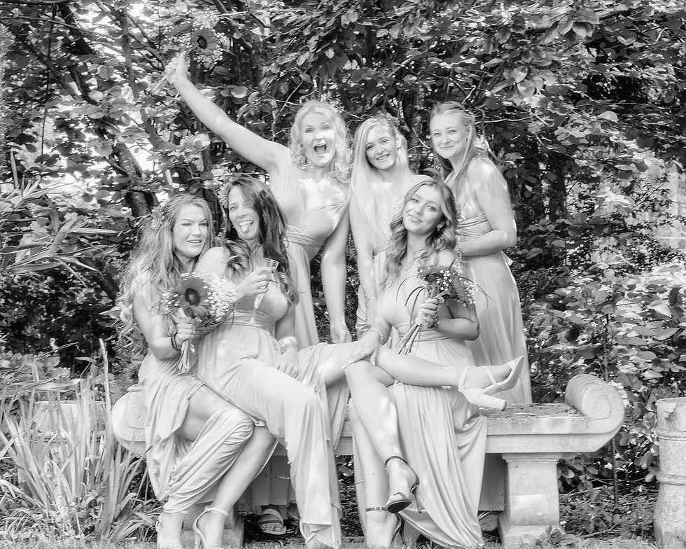 Six smiling women in dresses pose on a stone bench in a leafy garden, holding bouquets and laughing together.