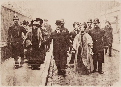 Sepia historical photo of women and policemen walking down a cobblestone street, all with serious expressions.