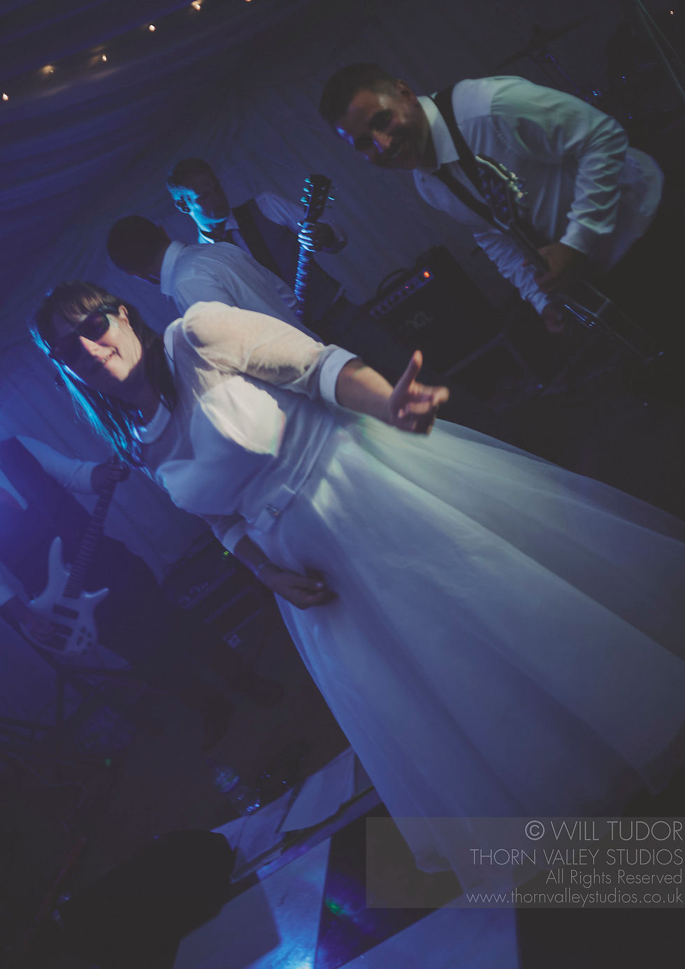 Band in white shirts and sunglasses performs under blue lights; woman in white dress smiles, guitars behind. © Will Tudor