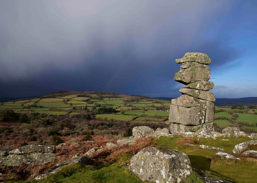 Stacked rock cairn on a hill above patchwork green fields, under a dramatic dark storm cloud sky.