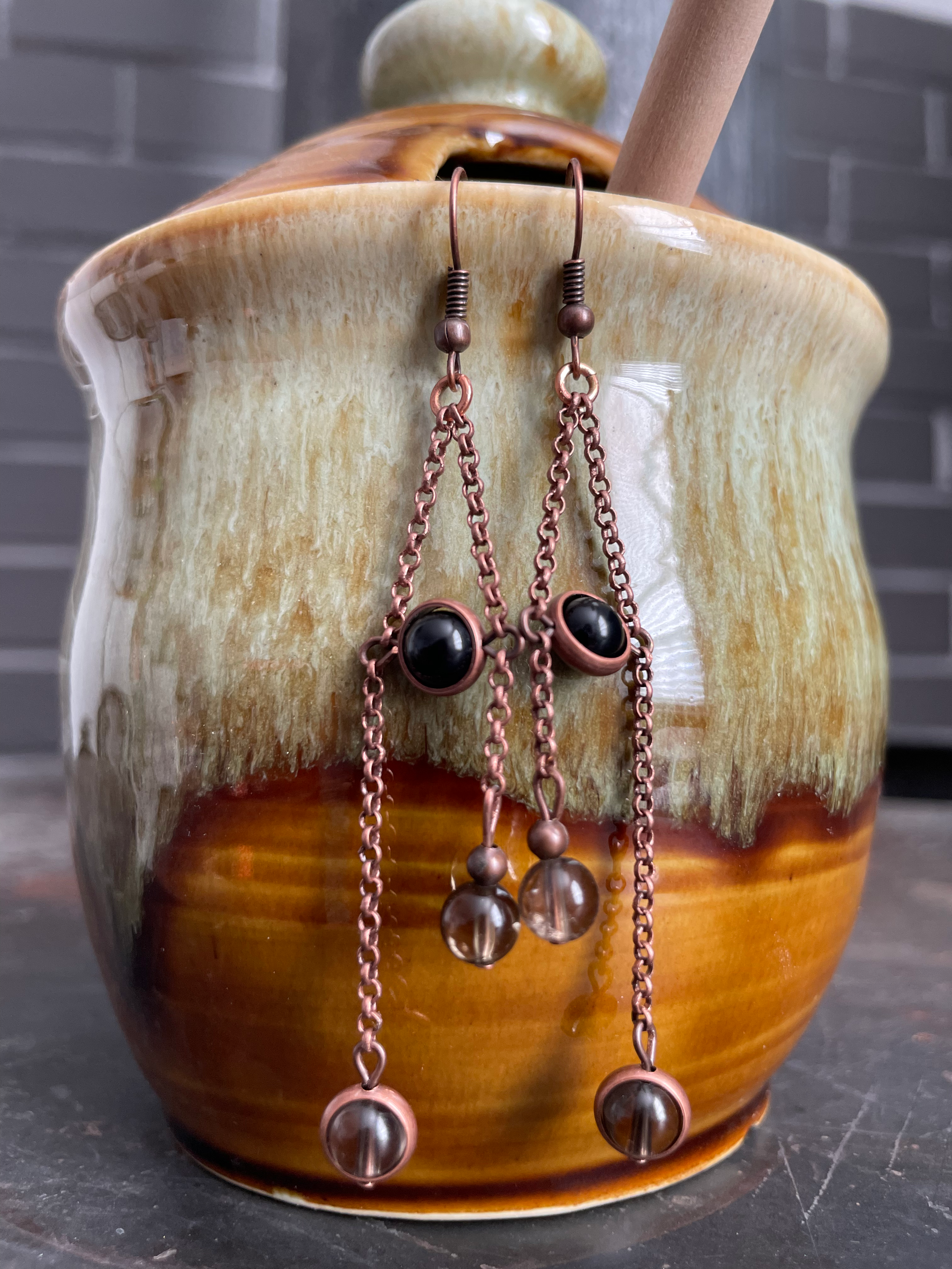 Copper earrings with onyx and smoky quartz dangle from a ceramic honey pot