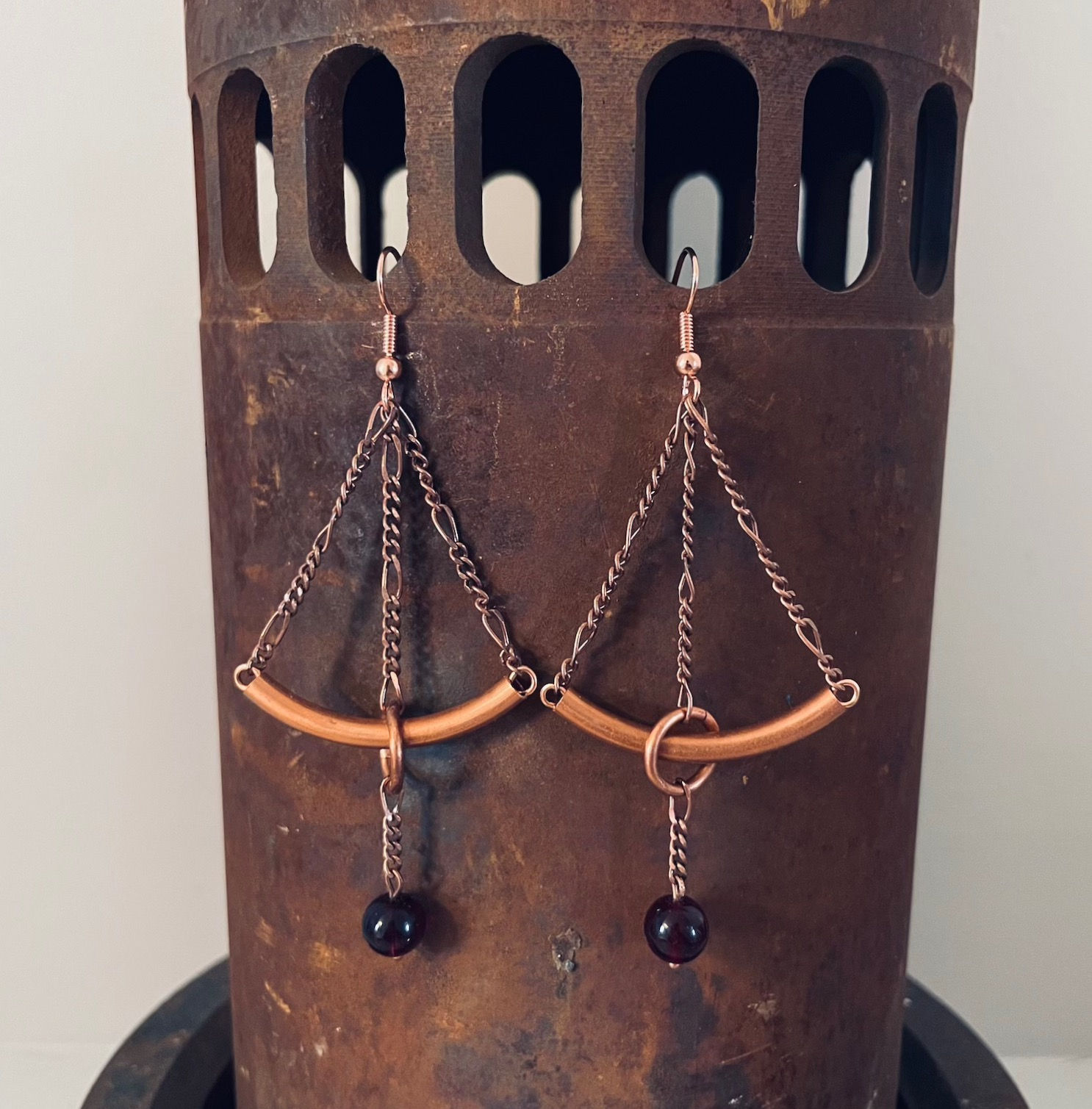Copper and ruby red glass earrings dangle from a rusted pipe