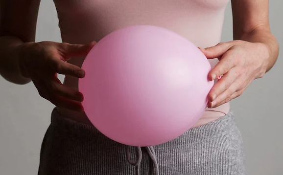 Person holding a pink balloon in front of their stomach, wearing a pink top and gray pants. Background is plain and neutral.