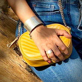 Woman wearing a wide silver cuff bracelet and ring holding a small mustard yellow quilted handbag on denim jeans