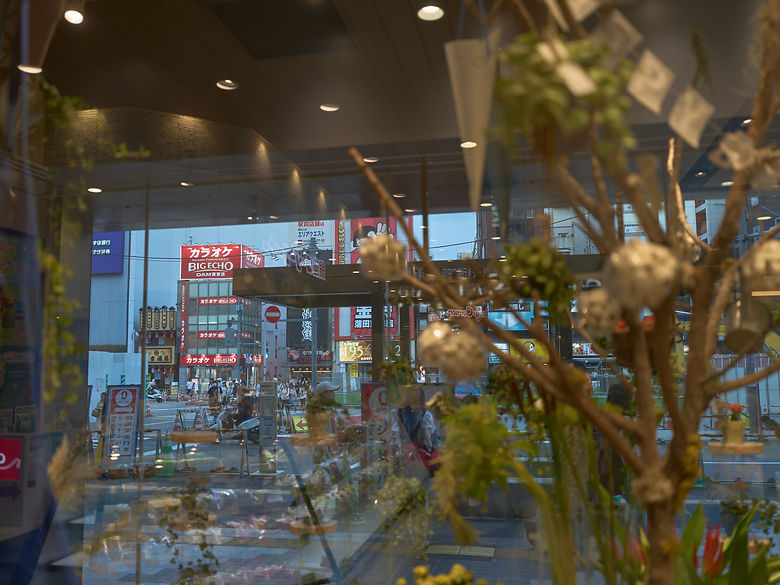 花や植物のある店内のガラス越しに、街の看板や通りと室内の照明が重なって見える風景。