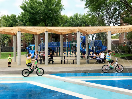 children riding bikes through a splash pad in front of a playground.