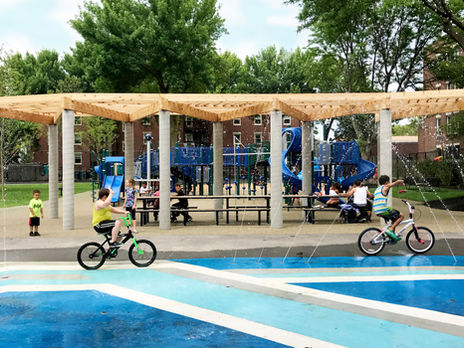 children riding bikes through a splash-pad in front of a playground.