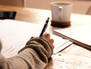 Close-up of a student’s hand writing in a notebook at a wooden desk, with a warm mug nearby — symbolising reflective growth, intentional planning, and the quiet discipline behind powerful portfolio-building.