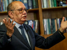 Ministro do STF Gilmar Mendes, de braços abertos, gesticulando e estante com livros ao fundo