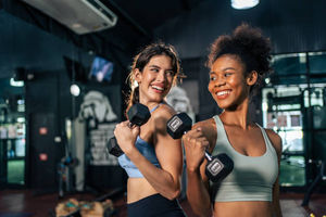Two women smiling while holding dumbbells in a public gym.