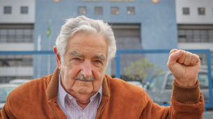 Foto de Pepe Mujica, ex-presidente do Uruguai, com uma blusa laranja erguendo o punho em saudação.