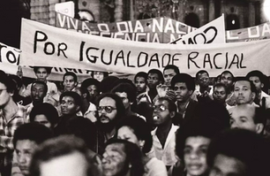 Fotografia histórica da Fundação do Movimento Negro Unificado, mostrando uma grande manifestação pública. No centro da imagem, uma faixa destaca a luta por igualdade racial, reforçando a importância do movimento na defesa dos direitos da população negra no Brasil. O ambiente é marcado por expressões determinadas e cartazes adicionais, evidenciando o engajamento dos participantes em promover mudanças sociais e combater o racismo estrutural.