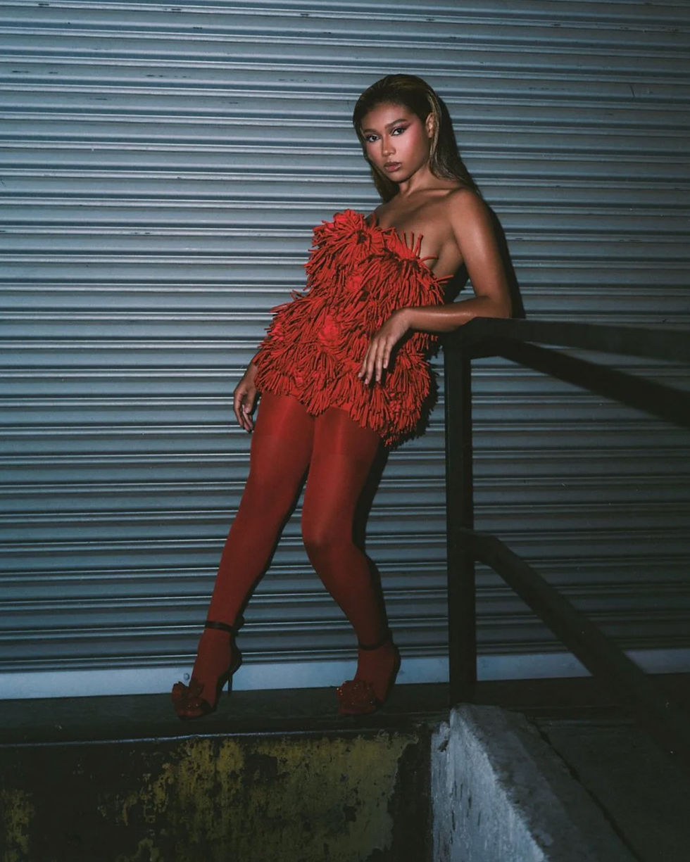 Sierra Capri leaning against a railing in Giseli Dias floral sandals and a textured red dress, photographed for Numéro Netherlands.