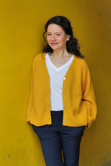 Schauspielerin Isabel Arlt vor gelbem Hintergrund. Mit Jeans, weißem T-Shirt und gelber Wollweste in lockerer Haltung
