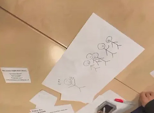 A simple black stick-figure drawing on a sheet of paper, lying on a table beside printed hint and vote cards.