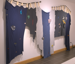 Long vertical strips of blue and grey fabric torn and stitched together to form a long, misshapen tapestry that resembles something between a crucified body and a waterfall caught in time. There are tears within each panel that have been mended with colorful fabric. The piece hangs from wide strips of canvas off of wonky, rusty, salvaged pipes from the river.
