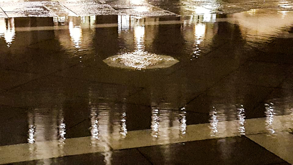 A night-time closeup of a puddle of water on a stone plaza. Lights are reflected in the water. In the center, a circular light spot shows where water is bubbling up from below the stones. 