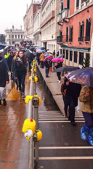 People with umbrellas traverse a raised boardwalk constructed next to shops. 