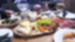 a table full of food, including an long oval wooden plate with cheese, grapes, apricots, and various meats. OlivA